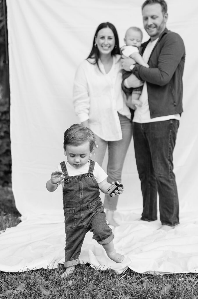 Toddler walking barefoot in front of the sheet backdrop while parents and baby smile behind him.