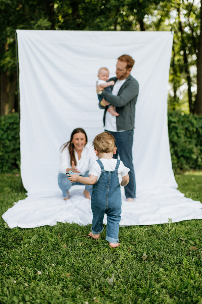 Young boy walking toward his mom while dad holds the baby in front of the white sheet backdrop.