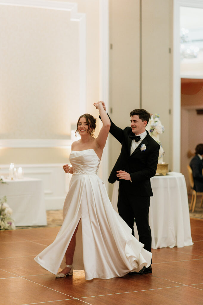 Groom twirls bride during their first dance in the Smith Ballroom with their wedding cake in the background.