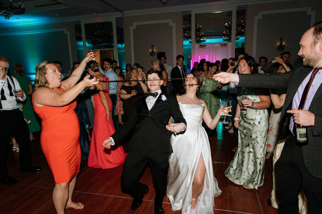 The bride and groom lean back together during a fun limbo game on the dance floor surrounded by cheering guests.