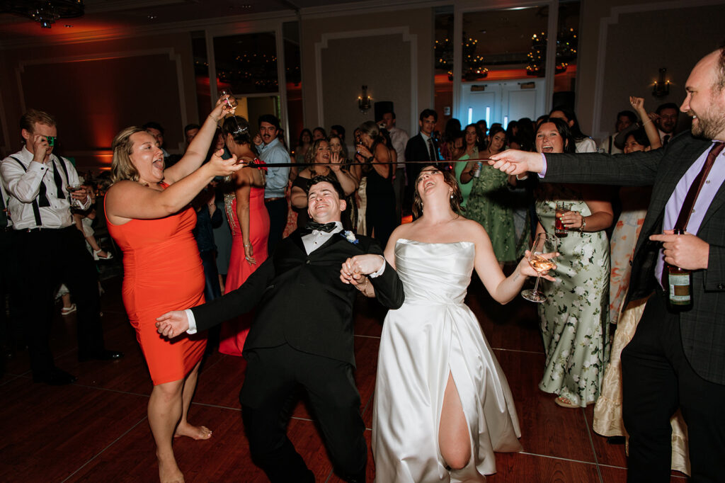 The bride and groom lean back together during a fun limbo game on the dance floor surrounded by cheering guests.