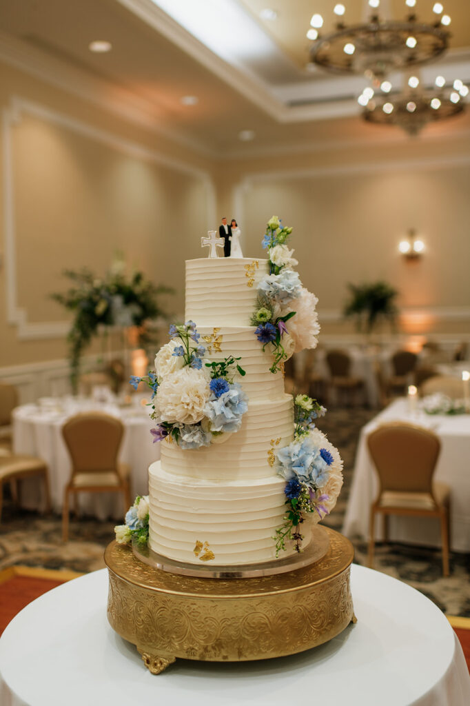 Three-tiered white wedding cake decorated with cascading pastel florals and a bride and groom topper, displayed in the Smith Ballroom.