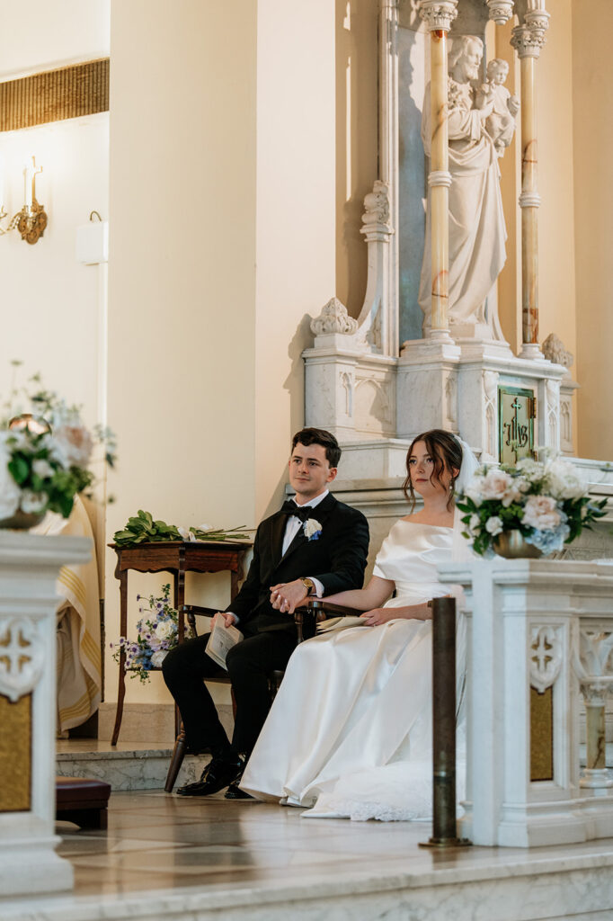 Bride and groom sit hand in hand, smiling during their Catholic mass at The Holy Spirit Chapel.