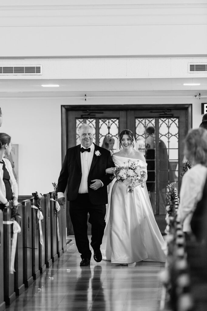 Bride walks arm in arm with her father down the aisle of The Holy Spirit Chapel at Saint Mary’s College.