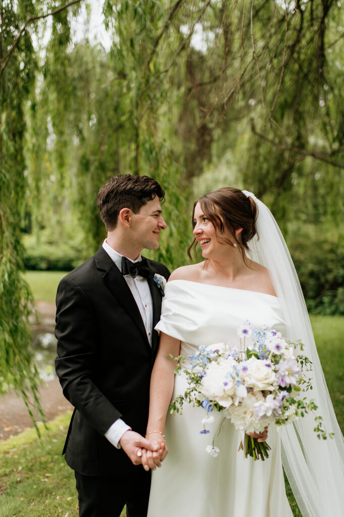 Bride and groom hold hands and laugh together under the willow trees near the water.