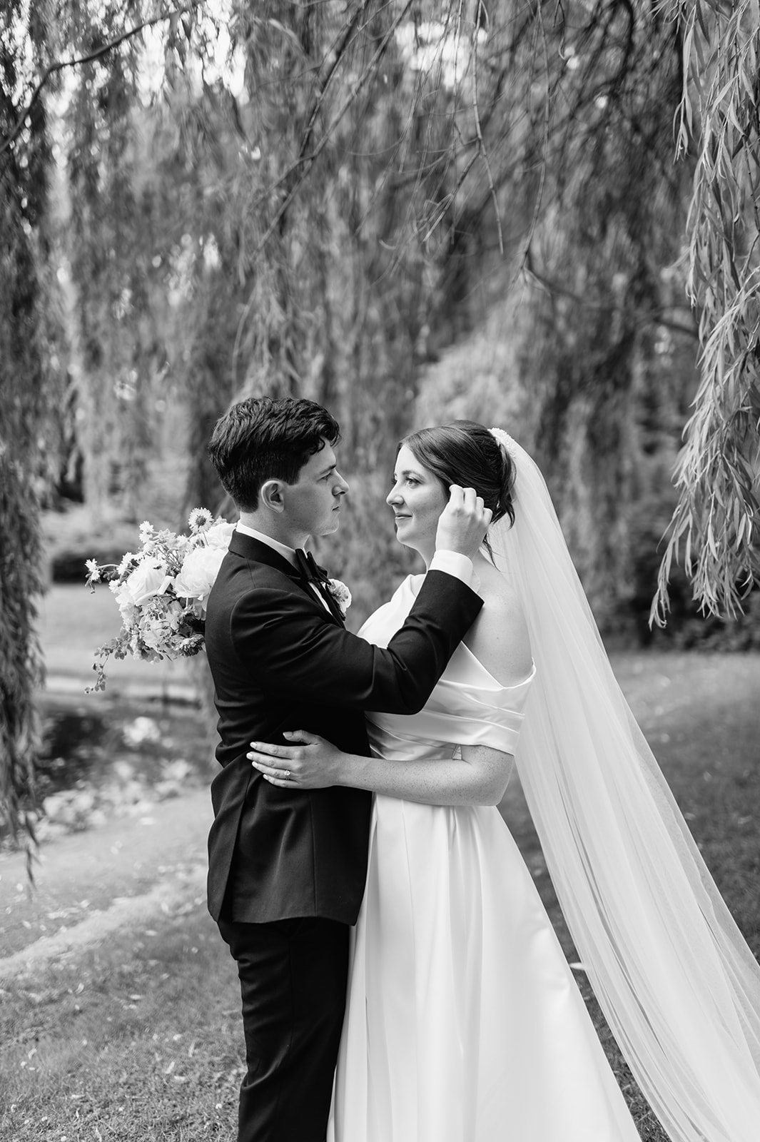 Black and white portrait of the groom brushing hair from the bride’s face during a quiet moment under the trees.