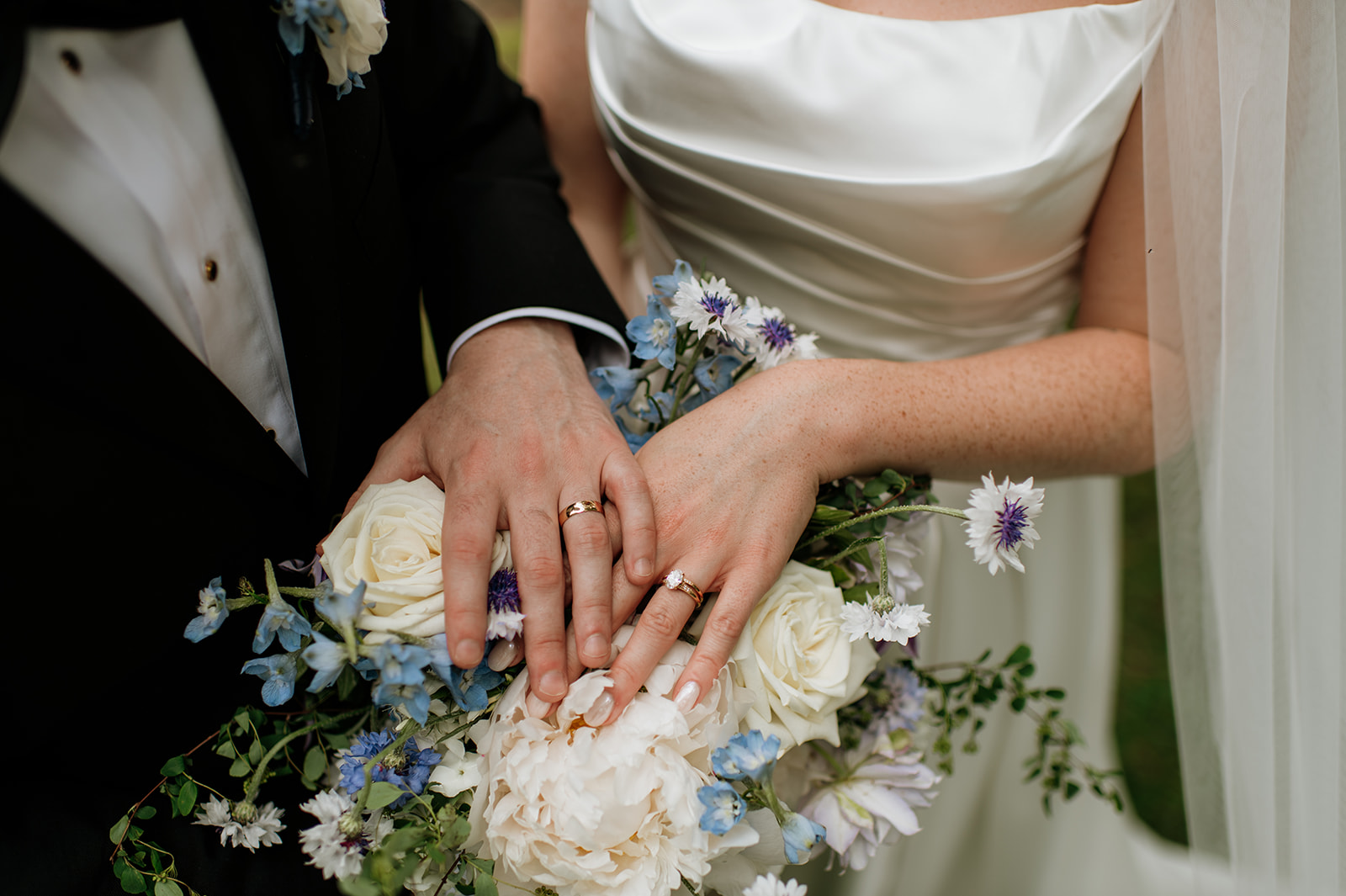 Close-up of bride and groom’s wedding bands resting on top of her bouquet of ivory and blue flowers.