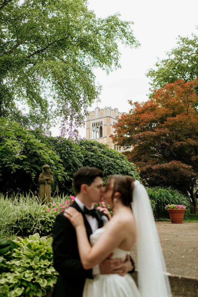 Blurred foreground of bride and groom kissing with a statue and Notre Dame campus building in focus behind them.