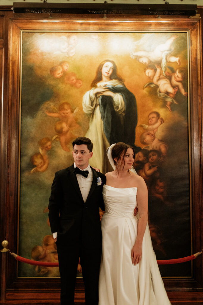 Bride and groom stand together in front of a large religious painting inside Notre Dame’s campus.