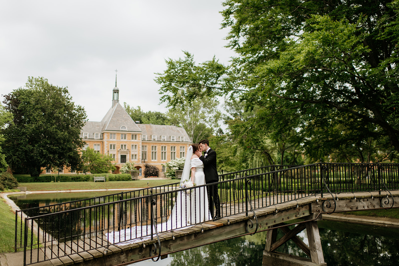 Bride and groom kiss on a small bridge with Saint Mary’s College building in the background.