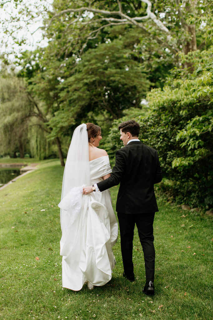 Bride and groom walk hand in hand along the pond at Saint Mary’s College, smiling at each other.