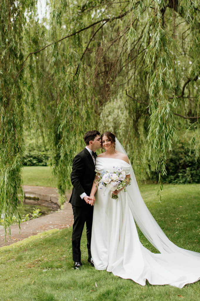Groom kisses bride’s temple as she holds her bouquet under sweeping willow trees at Notre Dame.