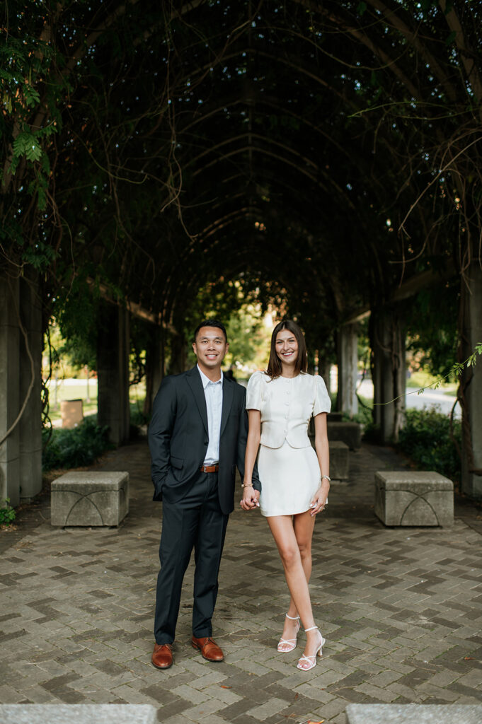 Couple standing hand in hand beneath the vine-covered walkway during elegant University of Notre Dame engagement photos.