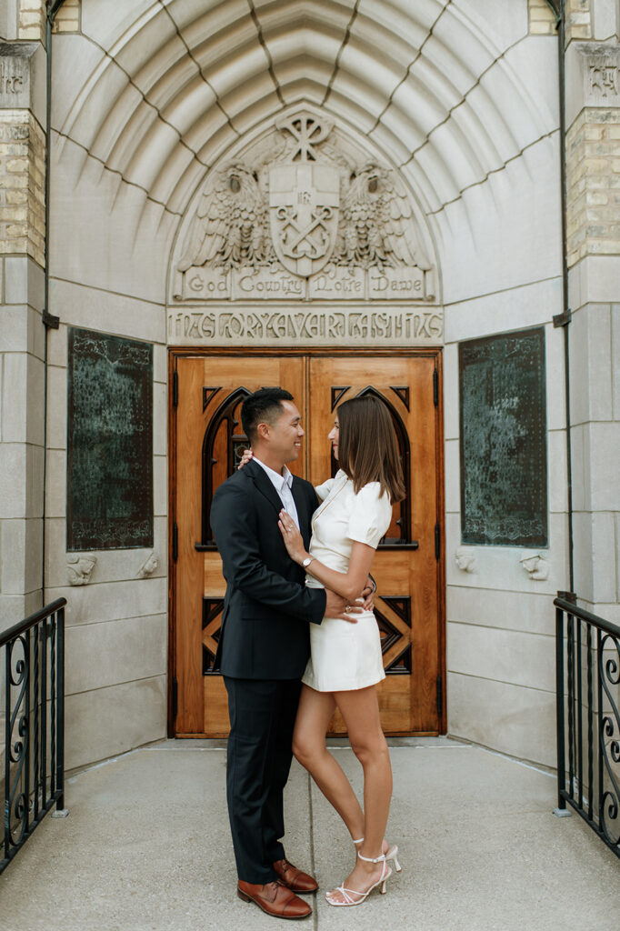 Couple posing outside of the Basilica of the Sacred Heart with the “God, Country, Notre Dame” doors behind them.