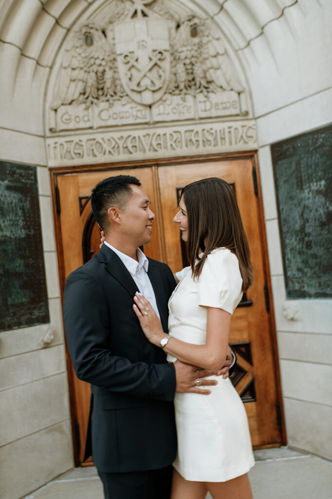 Candid photo of a couple posing outside of the Basilica of the Sacred Heart with the “God, Country, Notre Dame” doors behind them.