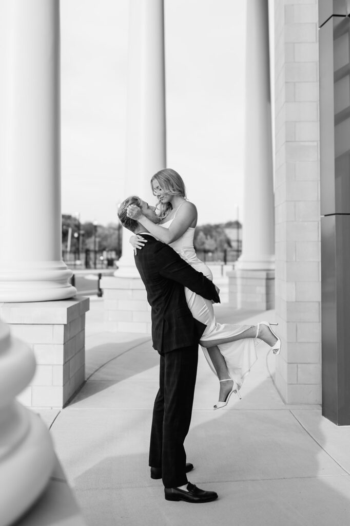 Black and white photo of a man lifting his fiancé near the grand pillars of the Elkhart County Courthouse