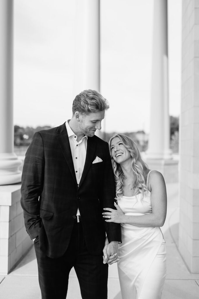 Black and white photo of a woman laughing up at her fiancé while holding hands in front of the courthouse columns in Goshen, Indiana