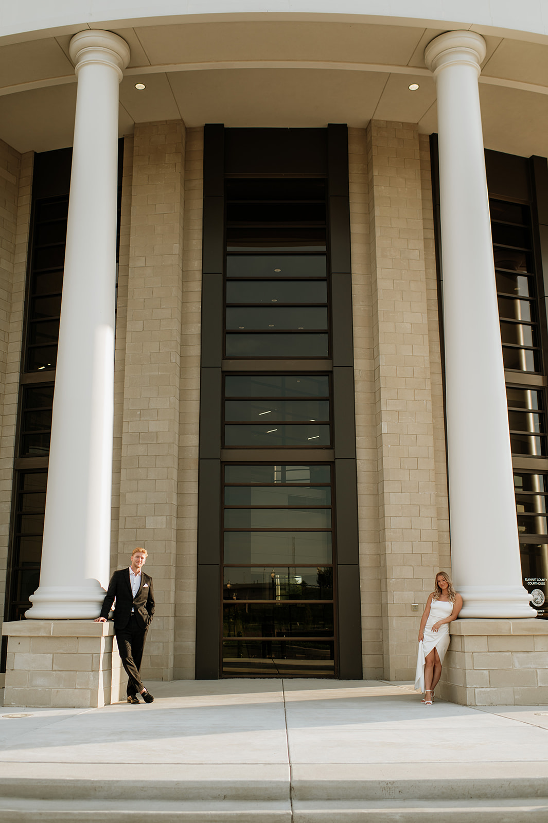 Wide photo showing the grand architecture of the Elkhart County Courthouse with the couple standing by opposite pillars