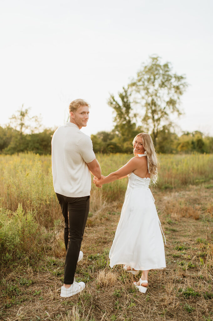 Couple looking back and smiling while holding hands in a field at sunset