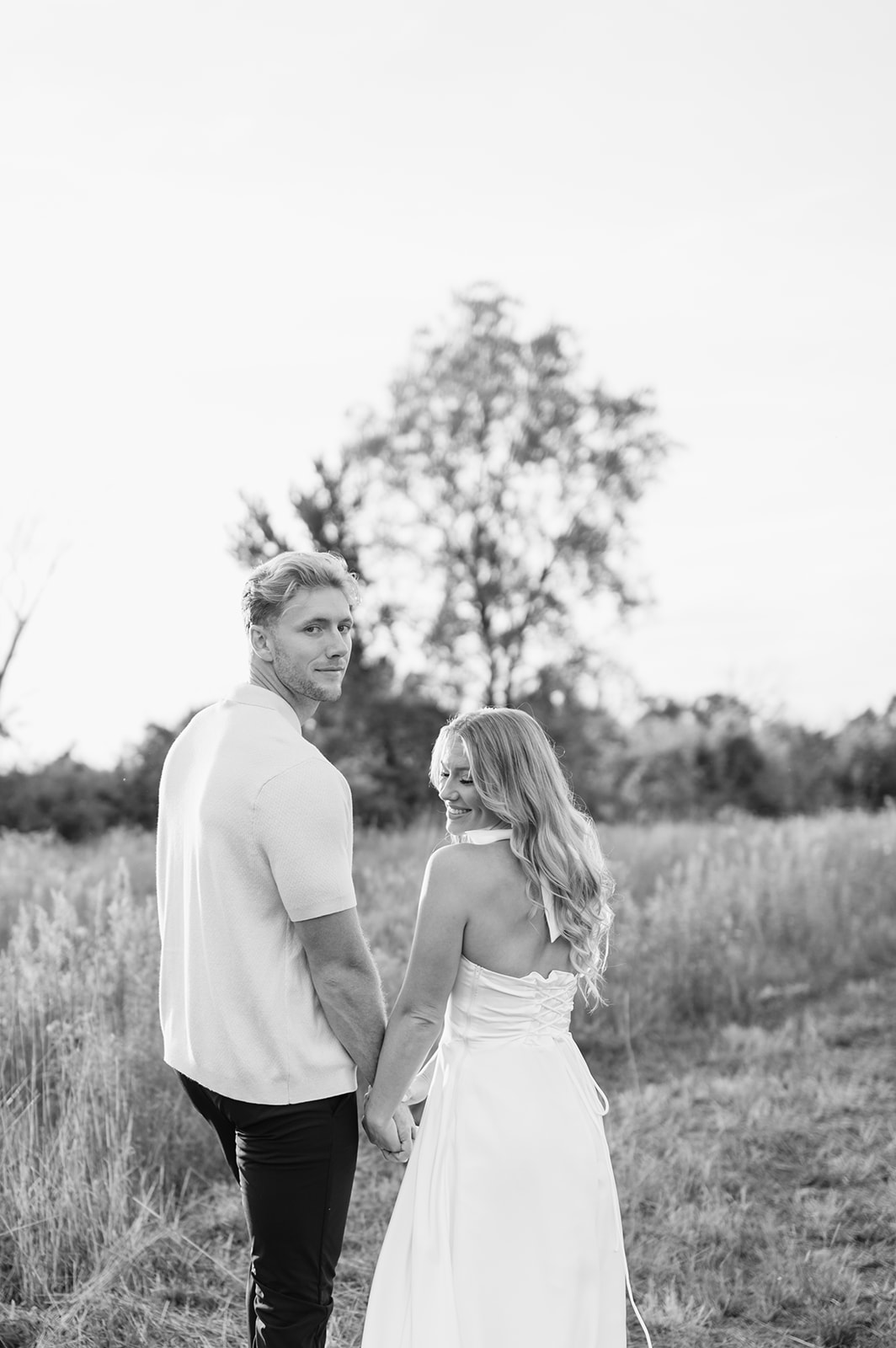 Black and white photo of a couple walking hand in hand through the field and looking back at the camera during sunset in Goshen, Indiana