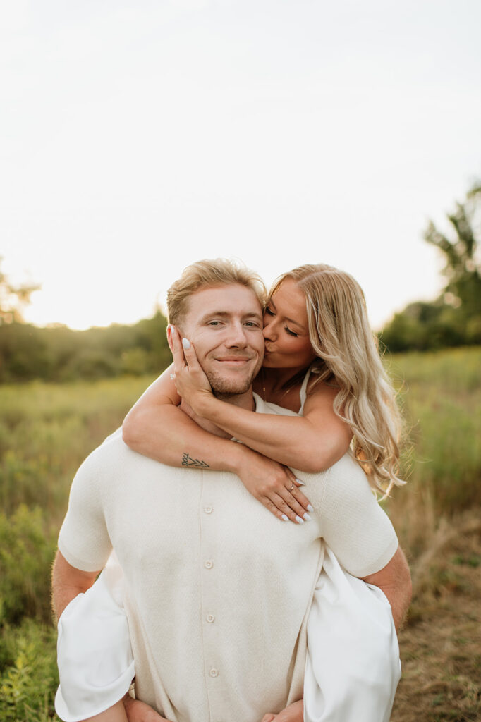 Woman playfully on man’s back kissing him on the cheek in the field during their Goshen, Indiana engagement photos