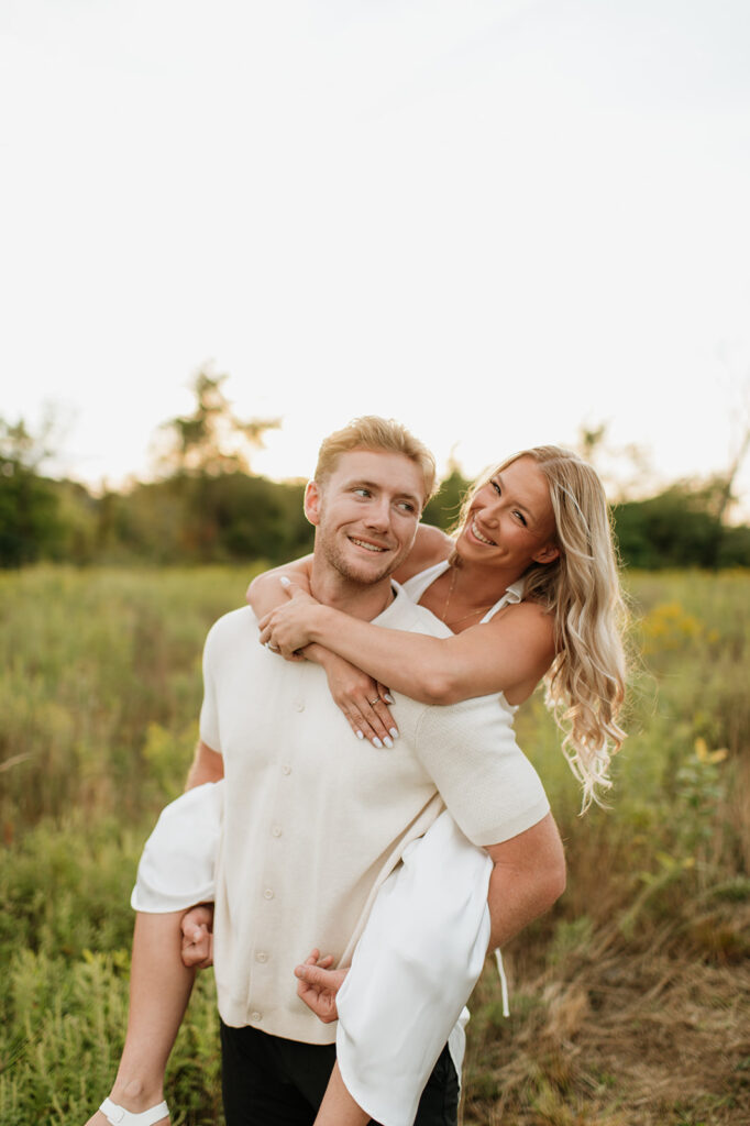 Woman playfully on man’s back as they laugh together in the field during their Goshen, Indiana engagement photos