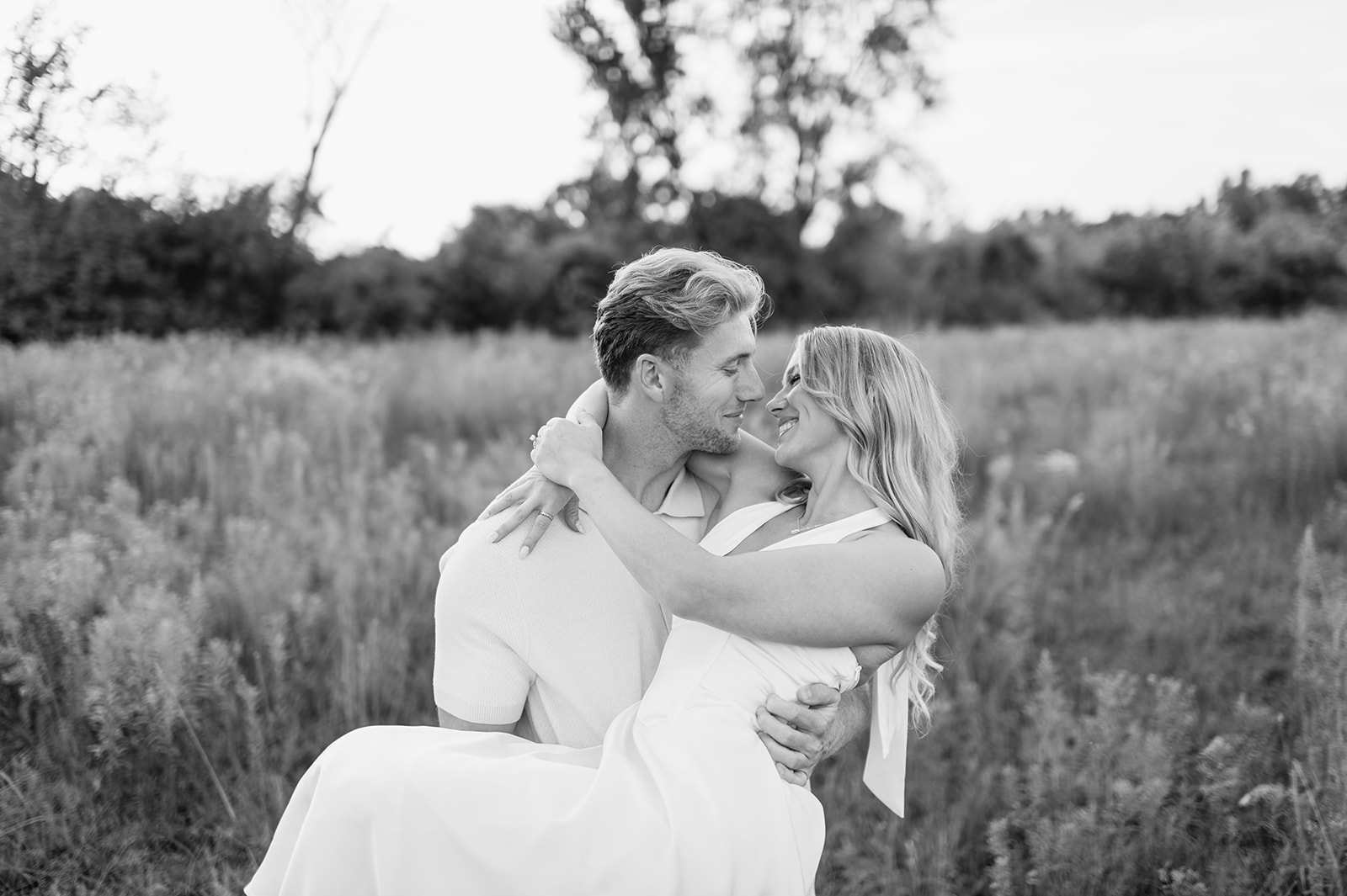 Black and white photo of a man carrying woman through the field as they smile at each other during their Goshen, Indiana engagement photos