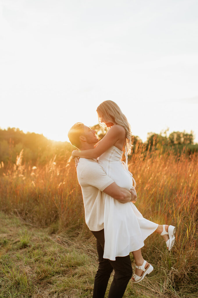Man lifting his fiancé during their Goshen, Indiana engagement photos in a field during golden hour