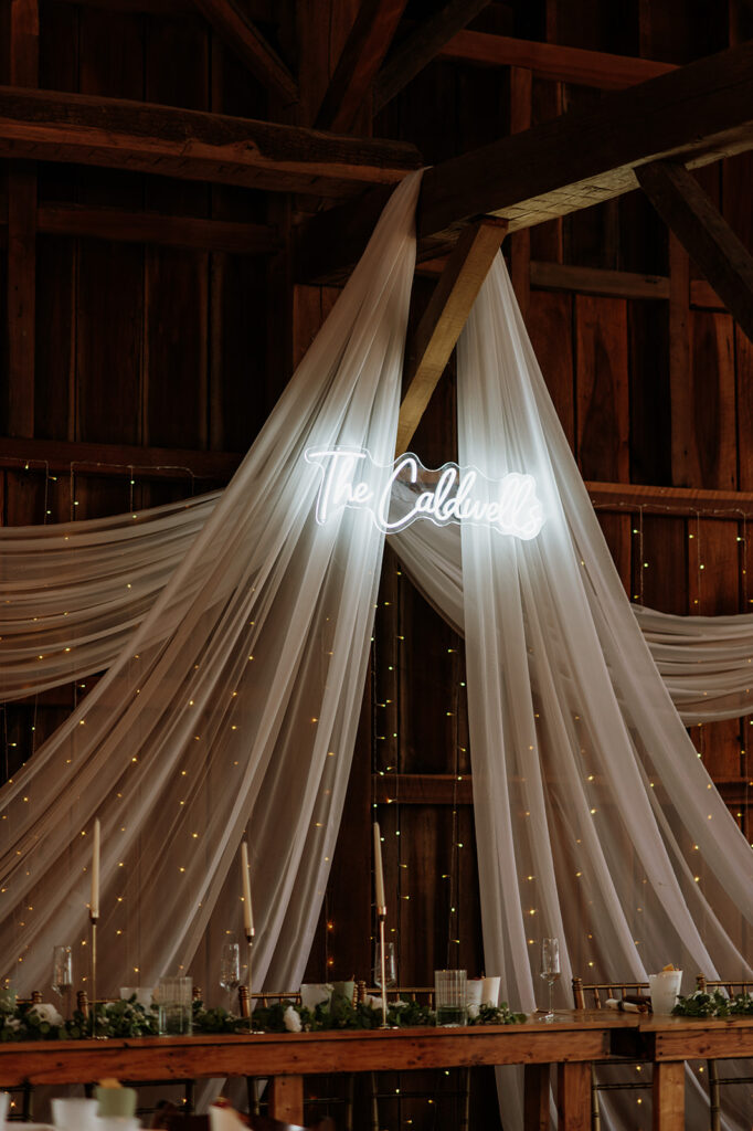 White neon “The Caldwells” wedding sign hanging above draped fabric and string lights inside the Event Barn at Rock Run Creek wedding reception.