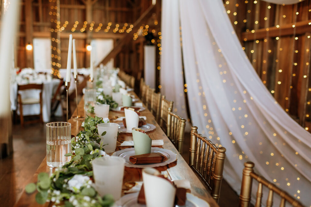 Long wooden head table decorated with candles, greenery, and custom guest cups at Rock Run Creek Barn wedding.