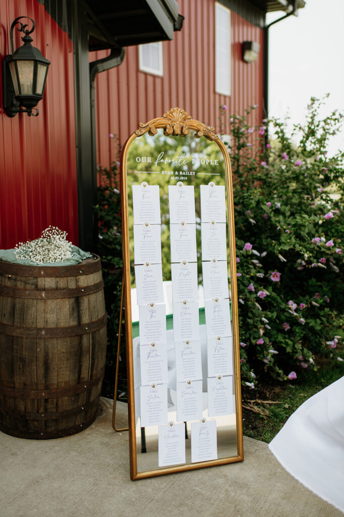 Gold-framed mirror seating chart for Ryan and Bailey’s Rock Run Creek Barn wedding guests displayed outside the red barn.