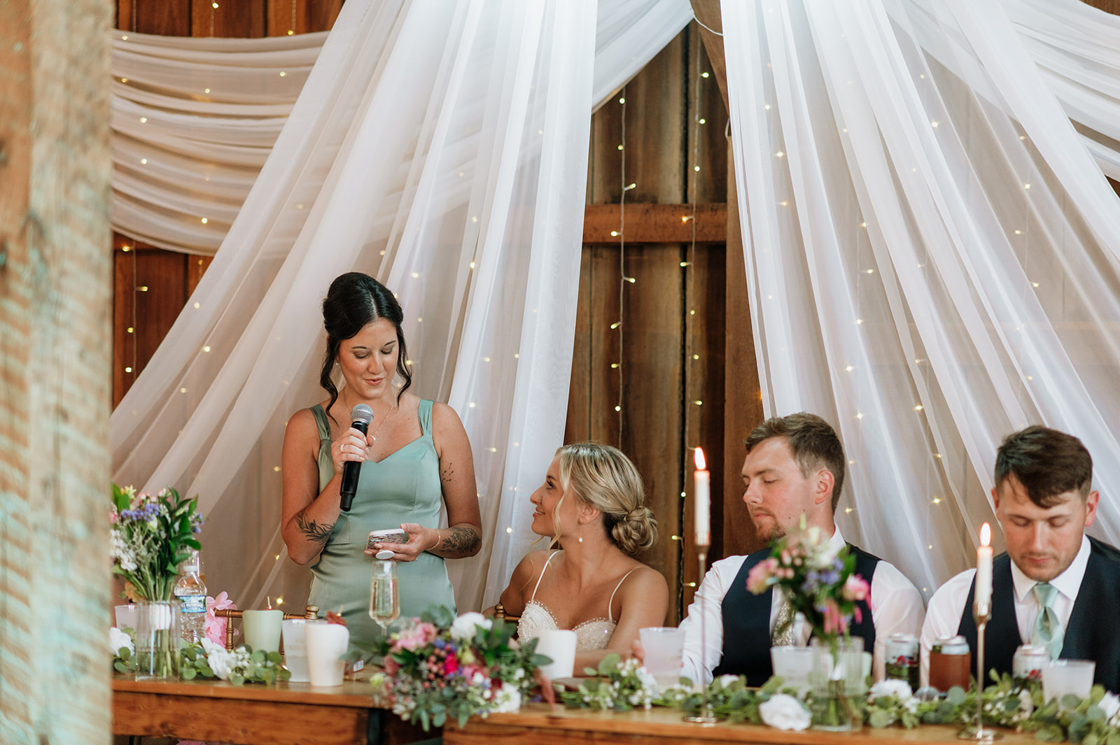 Maid of honor giving toast while bride smiles at head table during Rock Run Creek Barn wedding reception.