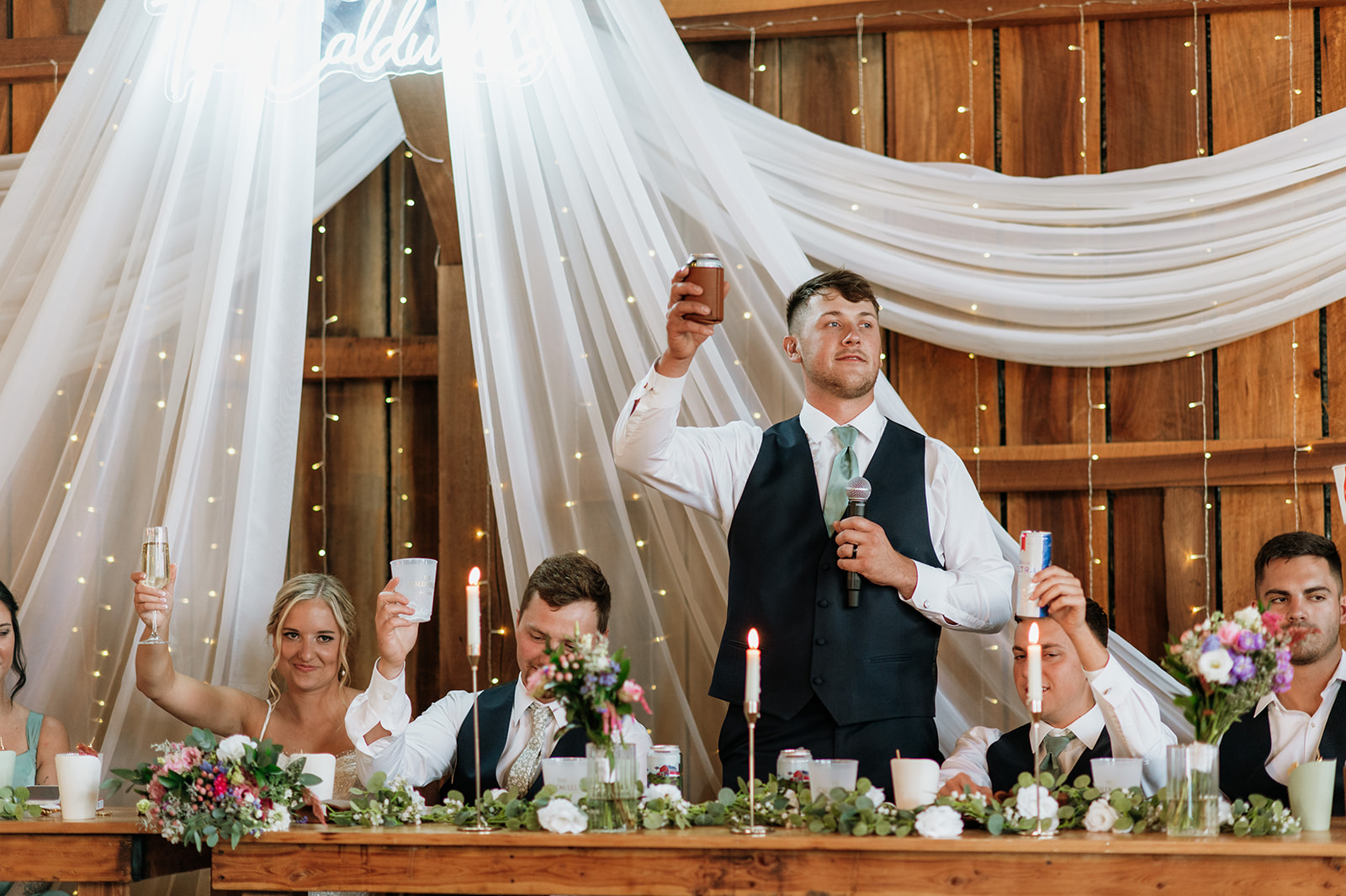 Best man raising glass for a toast as wedding party cheers at Rock Run Creek Barn reception.