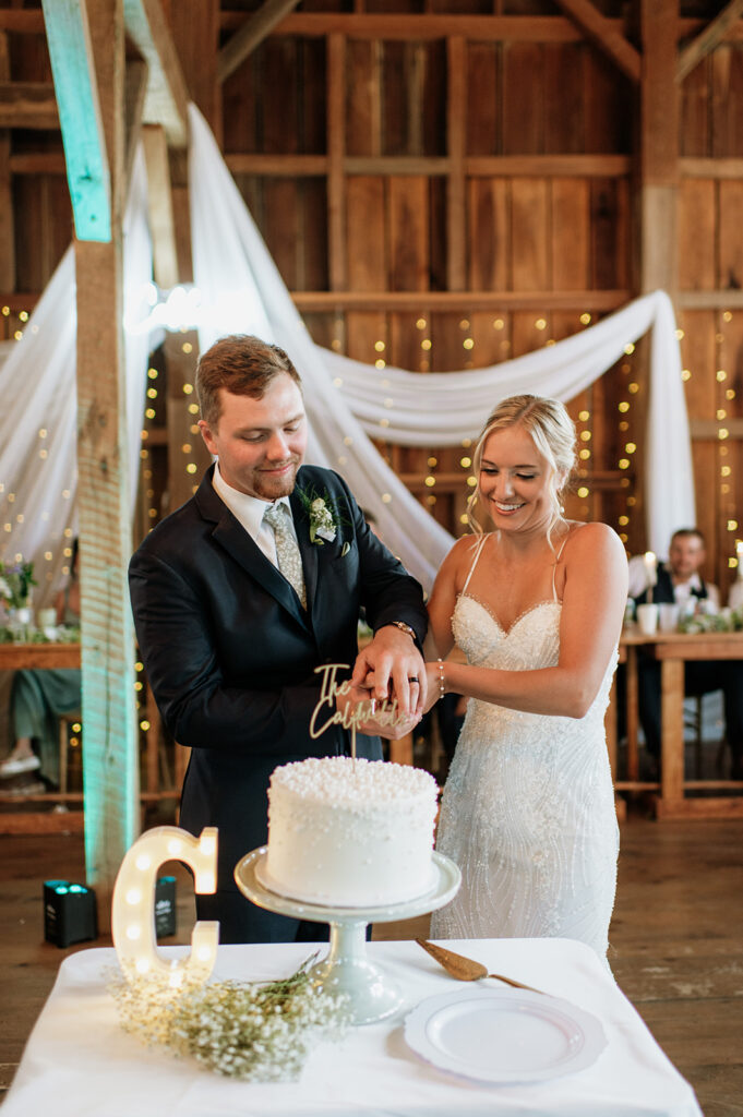 Bride and groom cutting their white wedding cake with “The Caldwells” topper inside Rock Run Creek Barn reception.