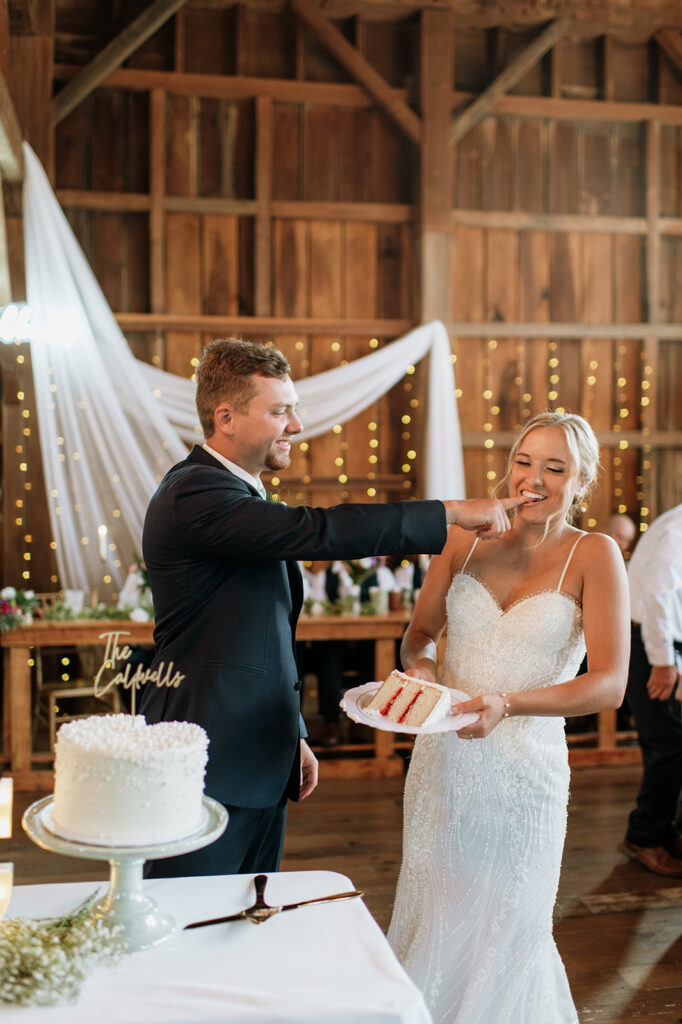 Groom feeding bride cake and laughing during reception at Rock Run Creek Barn in Goshen, Indiana.