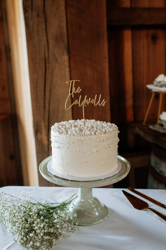 Simple white wedding cake with pearl details and custom “The Caldwells” topper displayed inside Rock Run Creek Barn reception.