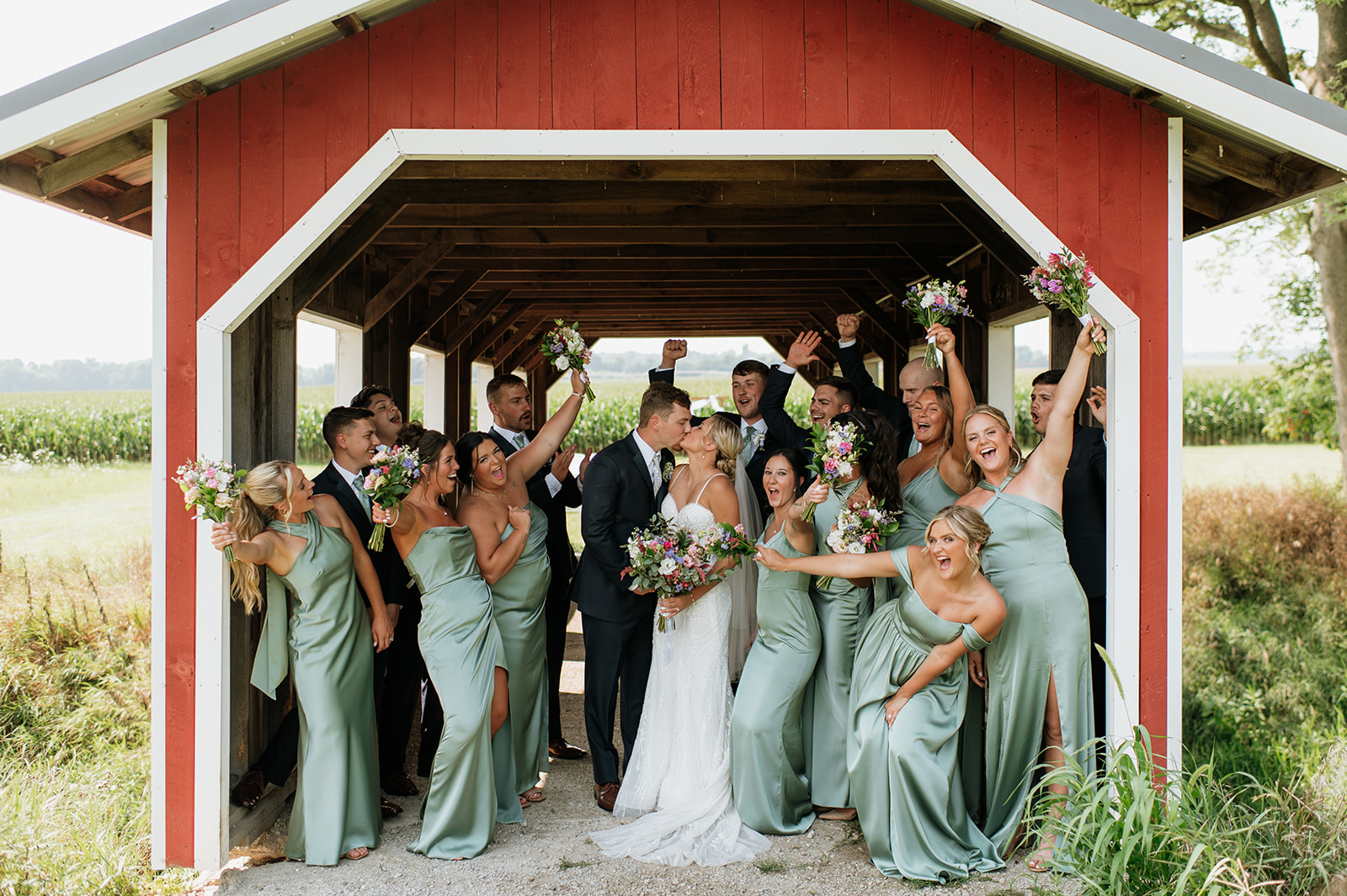 Bride and groom kissing surrounded by their wedding party underneath the covered bridge at Rock Run Creek Barn wedding venue