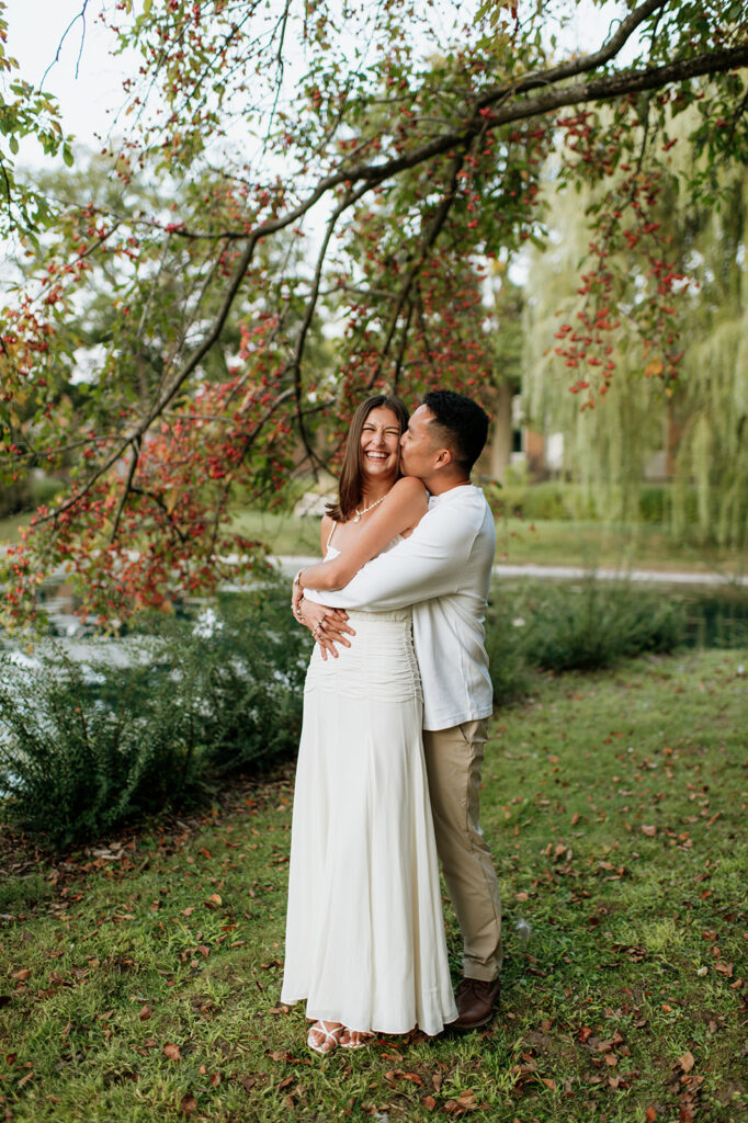 Man kissing his fiancé near Lake Marian surrounded by colorful trees during their Saint Mary’s College engagement photos.