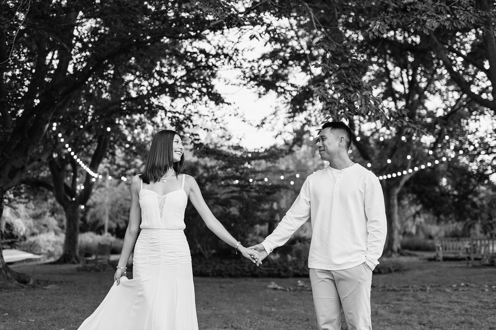 Black and white photo of a couple smiling and holding hands beneath trees wrapped in string lights at Saint Mary’s College.
