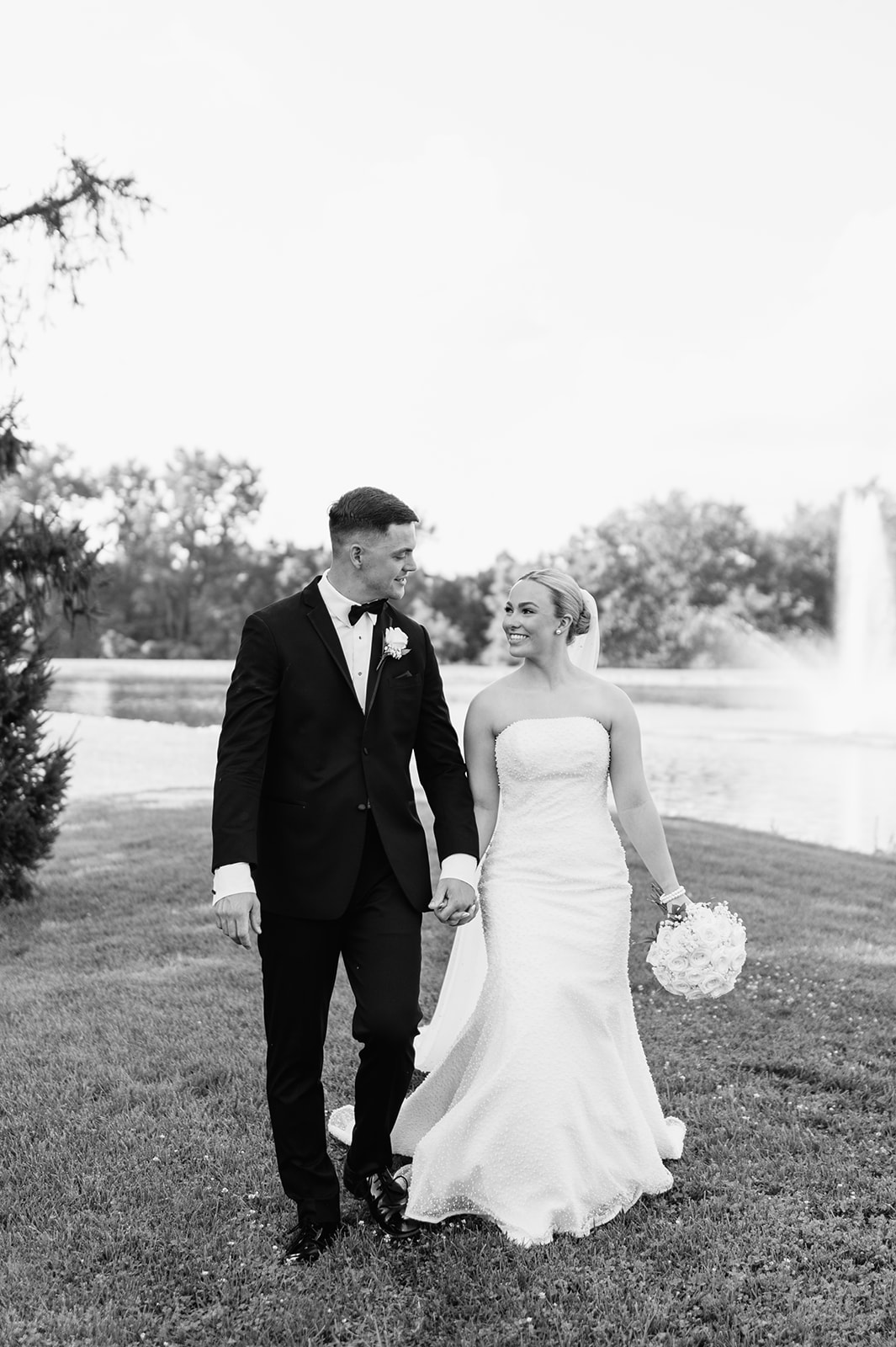 Black and white photo of a bride and groom walking hand in hand beside the pond at The Clubhouse venue during summer portraits.