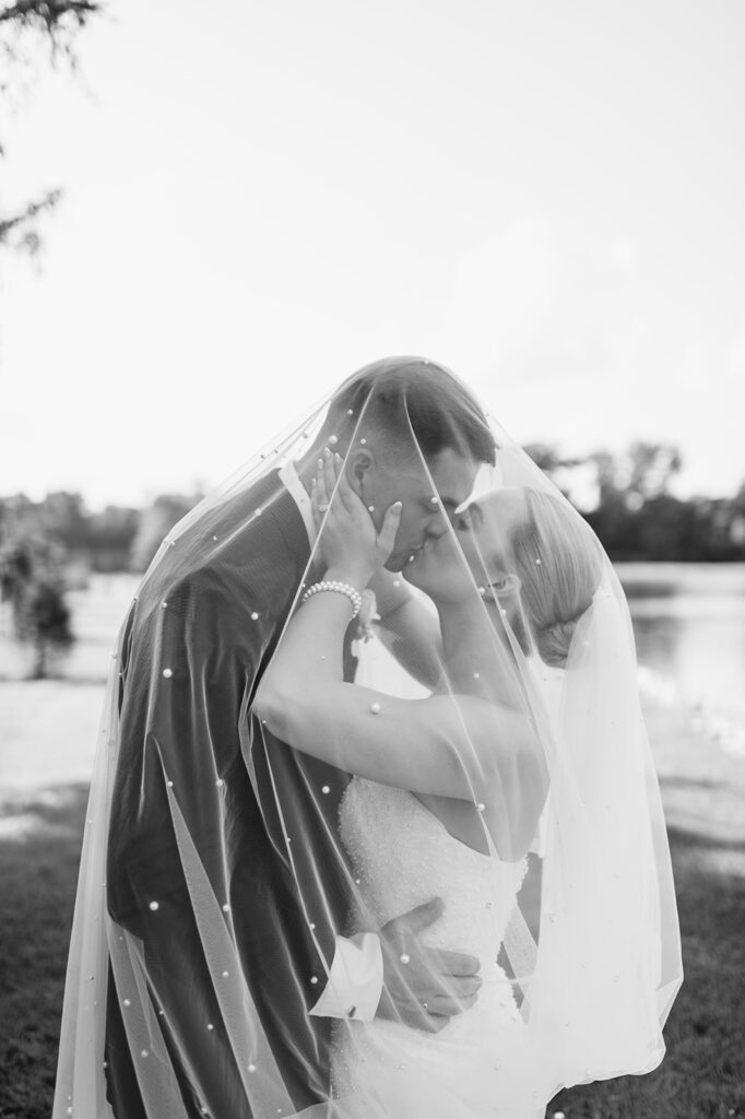 Black-and-white photo of bride and groom kissing under the bride’s veil on the grounds.