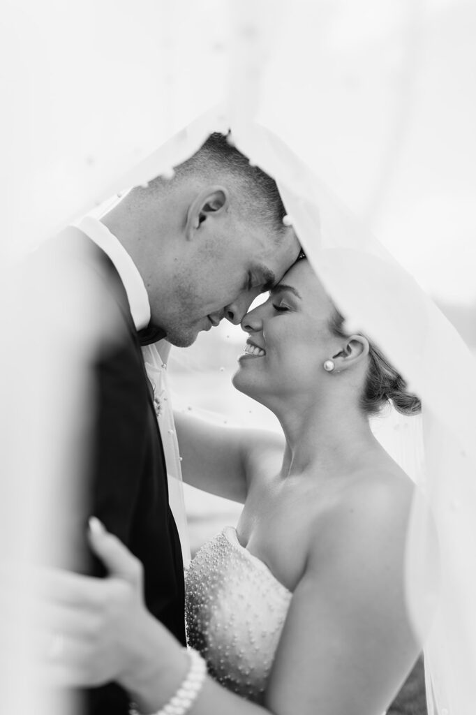 Black-and-white close-up of bride and groom forehead-to-forehead under veil at The Clubhouse venue.