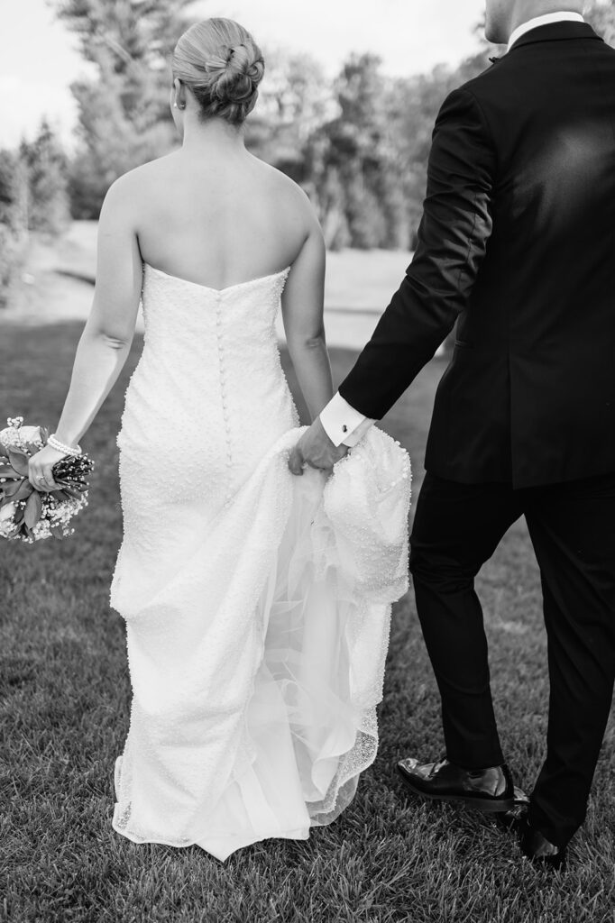 Black-and-white photo of couple walking away, groom holding the bride’s train.