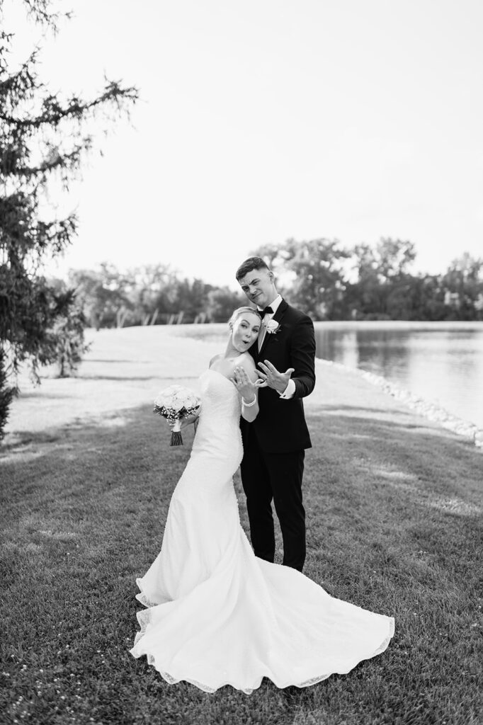 Black-and-white playful portrait of bride and groom showing their wedding rings by the water.