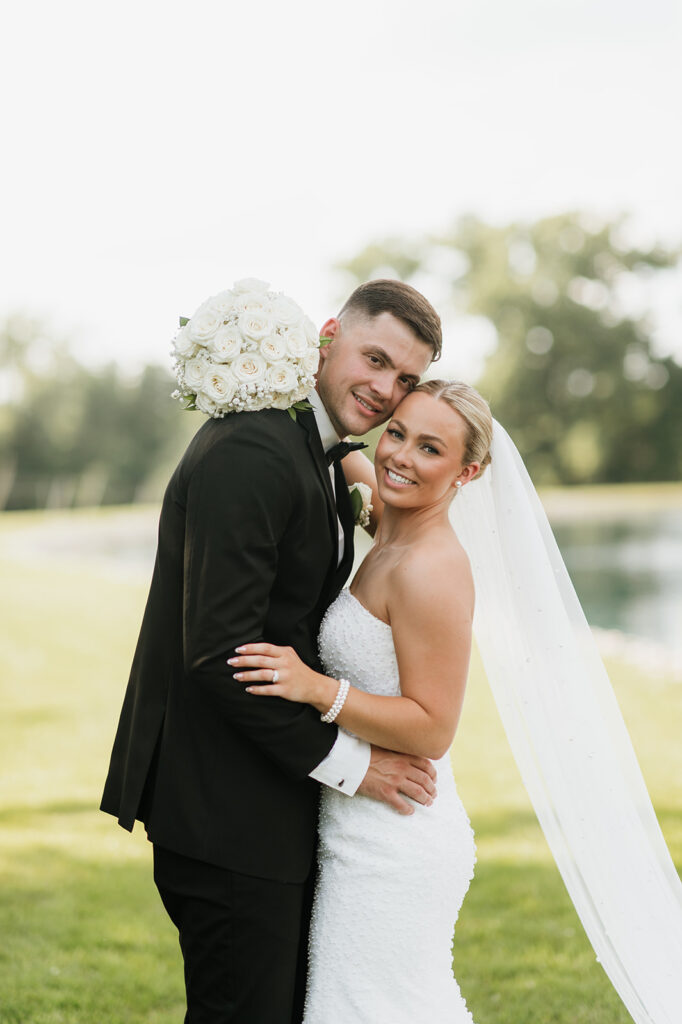 Groom hugging bride with her smiling toward camera, bouquet on his shoulder, taken on the grounds of The Clubhouse venue.