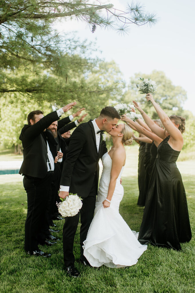 Bride and groom kissing under an arch made by the wedding party holding up bouquets at The Clubhouse Venue.