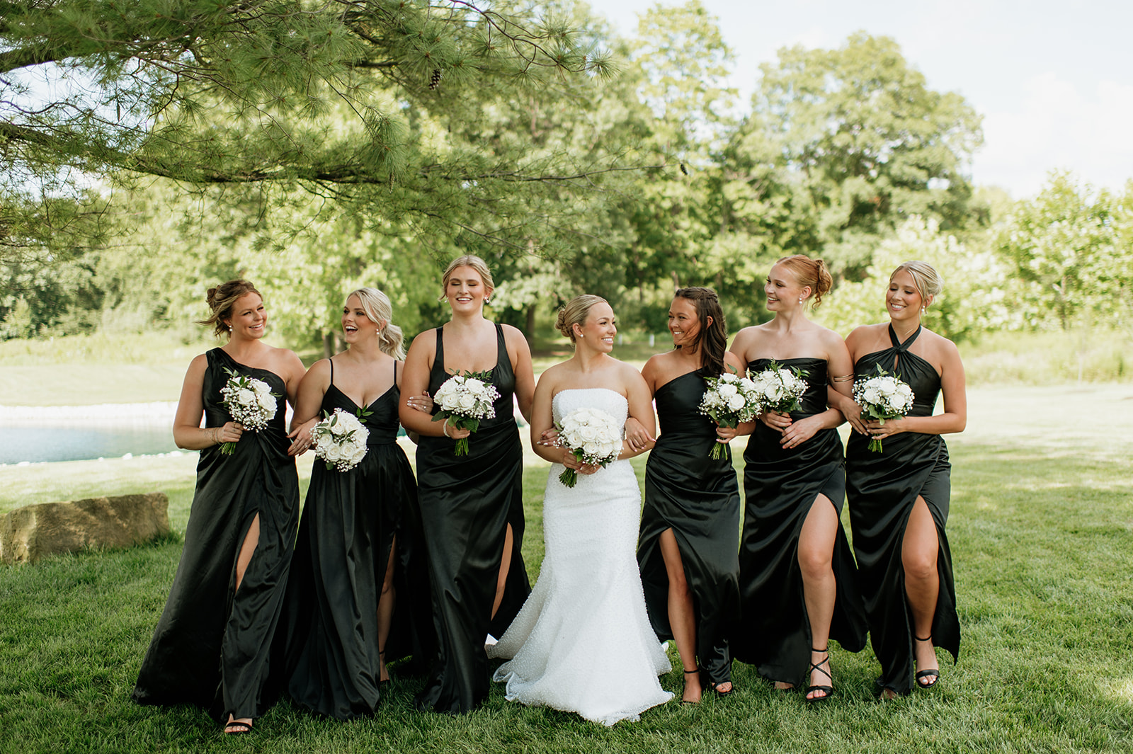 Bride walking arm-in-arm with her bridesmaids in black dresses on the grounds of The Clubhouse Venue.