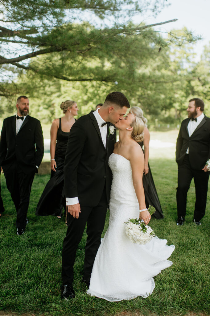 Bride and groom kissing with their wedding party around them on the lawn at The Clubhouse Venue.