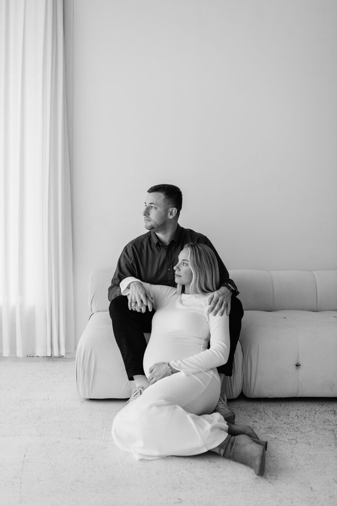Black and white photo of a couple seated together on a white couch during South Bend Indiana maternity photos at Area Hall Art House.