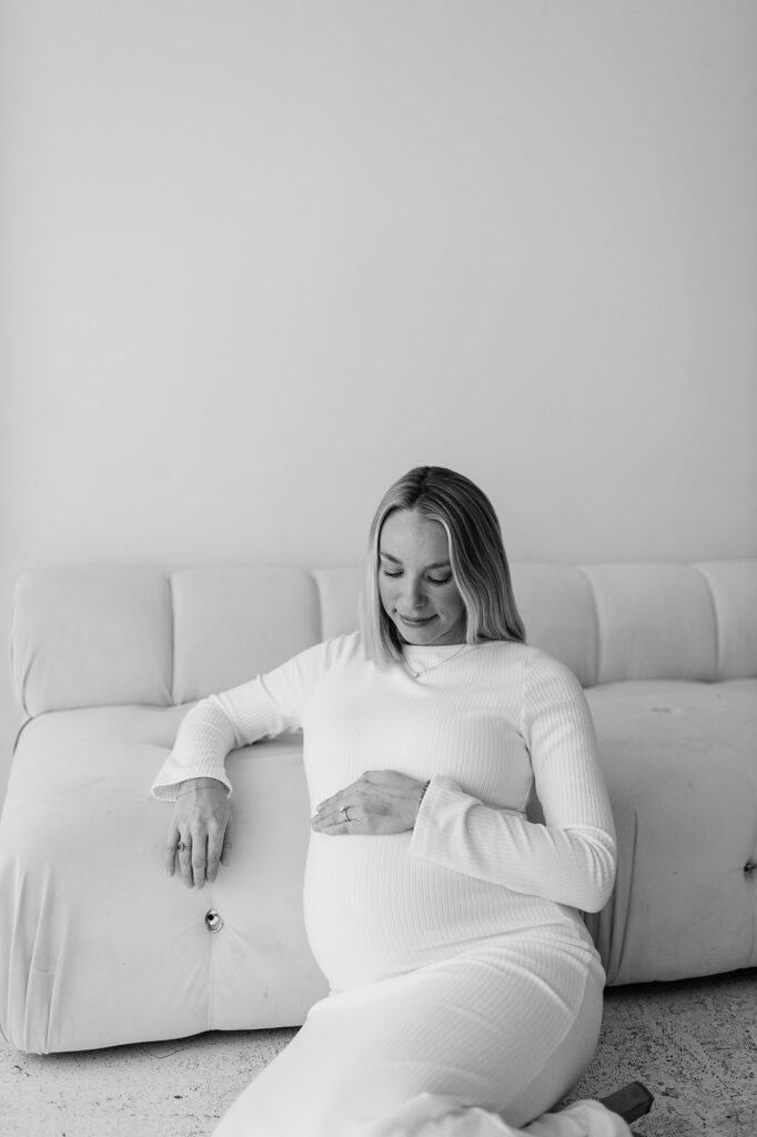 Black and white photo of a woman resting her hand on her belly while sitting against a couch at Area Hall Art House for her studio South Bend maternity photos.